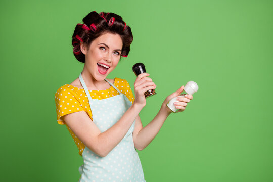 Photo of lovely charming funky young girl roller hairstyle hold pepper shaker salt bottle open mouth pretend having microphone sing wear dotted apron shirt isolated green color background