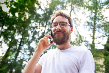Young smiling bearded man talking on a cell phone while standing outdoor on a sunny day. Using a mobile phone, smartphone.