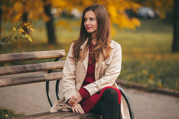 Portrait of young beautiful girl is wearing knitted dress and beige coat sitting on bench in the autumn park with yellow maple leaves.
