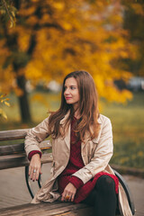 Fototapeta na wymiar Portrait of young beautiful girl is wearing knitted dress and beige coat sitting on bench in the autumn park with yellow maple leaves.