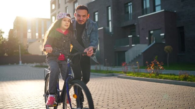 Dad is teaching daughter how to ride bicycle at sunset. Slow motion