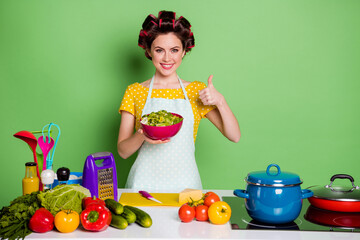 Photo model girl housewife cook supper kitchen table tomato cucumber prepare bowl eco salad show thumbup symbol wear hair rollers trendy yellow dotted dress isolated green color background