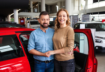 Portrait of smiling adult couple standing in front of a vehicle after buying a new car at dealership.
