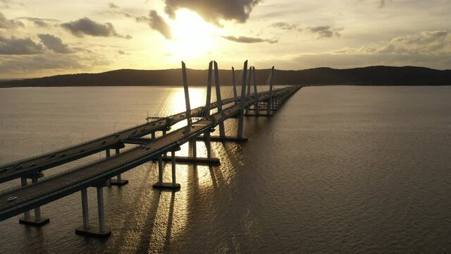 An aerial shot along the north side of the Mario M. Cuomo Bridge during a golden sunset. The drone camera dolly out over the Hudson River flying away from South Nyack, NY in the background.