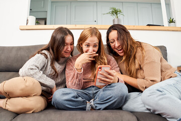 Group of female friends on the sofa at home, using a mobile phone laughing.
