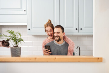 Happy young couple in kitchen at home, using mobile phone and laughing.