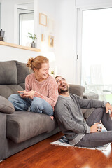 Happy young couple laughing holding cups of coffee and using phone on the couch together in the living room.