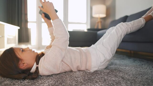 Little girl using digital tablet lying on her back on the floor in a cozy room