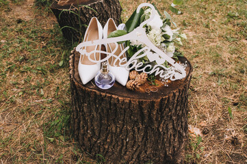 Wedding accessories and details of the bride close-up, top view: white hanger, leather shoes, bouquet, rings, cones, hairpin, perfume. Female set on a wooden stump, photography, concept.