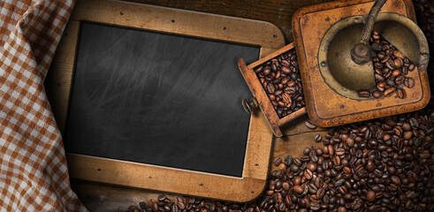 Old manual coffee grinder made of metal and wood with roasted coffee beans on the table with an empty blackboard and a checkered tablecloth.