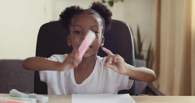 Webcam view of small mixed race girl speaks online via conference call chat, shoots video for blog, shows marker draws picture at table on paper, child learns remotely from home, e-learning concept