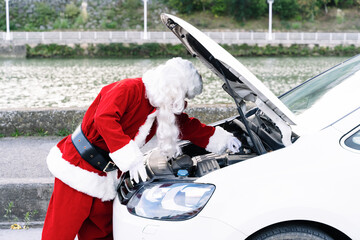 Portrait of Santa Claus with the car bonnet open and touching the engine because his car has broken down and he is wearing a face mask due to the coronavirus covid19 pandemic in Christmas 2020