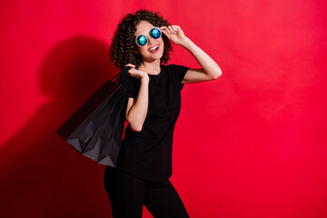Photo of cute sweet curly woman wear casual black outfit arm eyeglasses holding bags back isolated red color background