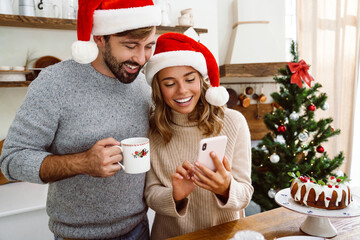 Charming cheerful couple in Christmas hats smiling and using cellphone