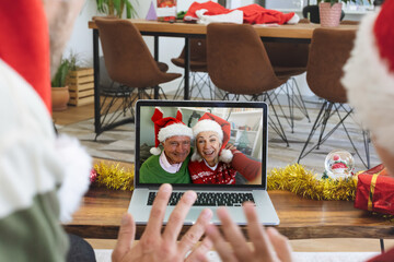 Fototapeta na wymiar Rear view of man and son in santa hats waving while having a videocall with senior couple in santa h
