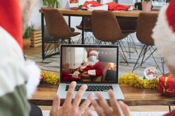 Fototapeta na wymiar Rear view of man and son in santa hats waving while having a videocall with santa claus wearing face