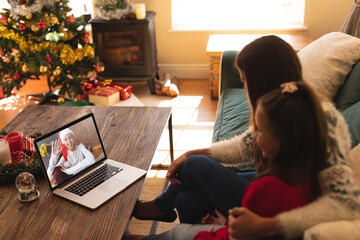 Woman and daughter sitting on couch having a videocall with woman in santa hat waving on laptop at h