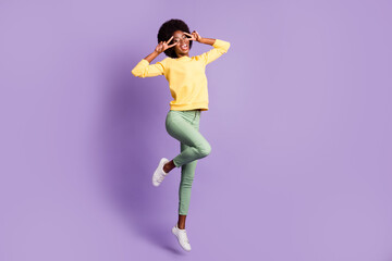 Full length photo of carefree dark skin girl jump make v-sign wear yellow sweater green pants isolated on violet color background