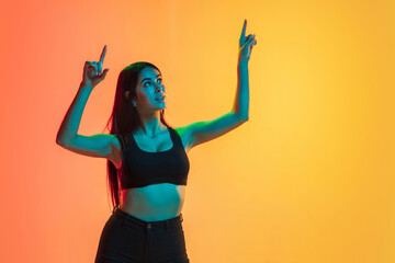 Pointing up. Young caucasian woman's portrait on gradient yellow-orange studio background in neon light. Concept of youth, human emotions, facial expression, sales, ad. Sportive fit brunette model.