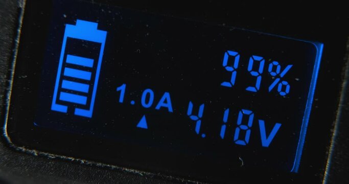 Screen of digital charging device with glowing blue percent digits and battery cell picture on black background extreme closeup