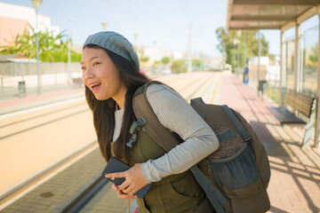 outdoors portrait of young happy and beautiful Asian Chinese tourist woman with backpack holding mobile phone on train station waiting excited in holidays travel and vacation