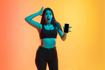Showing phone's screen. Young caucasian woman's portrait on gradient yellow-orange studio background in neon light. Concept of youth, human emotions, facial expression, sales, ad. Sportive fit model.