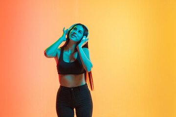 Listening to music in headphones. Young caucasian woman's portrait on gradient yellow-orange studio background in neon light. Concept of youth, human emotions, facial expression, sales, ad. Copyspace