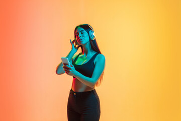 Listening to music in headphones. Young caucasian woman's portrait on gradient yellow-orange studio background in neon light. Concept of youth, human emotions, facial expression, sales, ad. Copyspace