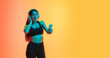 Listening to music in headphones. Young caucasian woman's portrait on gradient yellow-orange studio background in neon light. Concept of youth, human emotions, facial expression, sales, ad. Flyer