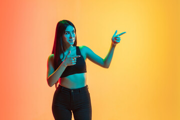 Pointing at sides. Young caucasian woman's portrait on gradient yellow-orange studio background in neon light. Concept of youth, human emotions, facial expression, sales, ad. Copyspace for ad.
