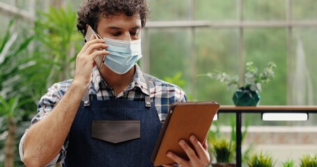 Fototapeta premium Close up portrait of young Caucasian male worker of flower shop typing on tablet and calling on smartphone. Handsome man garden center manager in mask tapping on device and talking on cellphone