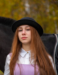 Red-haired girl in a black hat with a horse in the autumn forest.