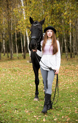Red-haired girl in a black hat with a horse in the autumn forest.