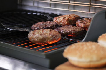 The process of cooking beef patties for burgers on a gas grill. Turkey and Beef cutlets cooked on a gas grill outside. In the foreground Burger buns with sesame seeds