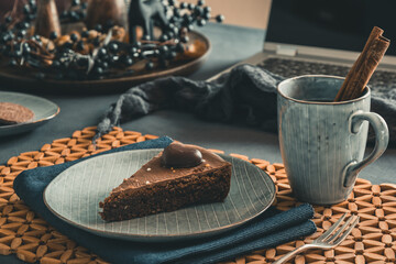 Piece of homemade chocolate pie and a cup with tea and cinnamon stick on a grey plate on a autumnal decorated table, open laptop in the background.