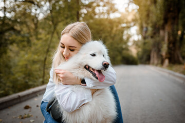 woman in white shirt is hugging her white dog samoyed outdoors in the park.