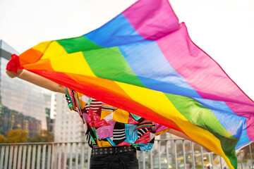 Back view of a person holding rainbow flag on city street
