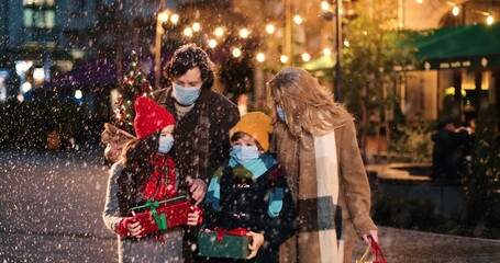 Portrait of Caucasian happy family walking with presents outdoors. Joyful kids with parents on street in city with little new year tree while snowing. Holiday lights and decoration. Christmas concept