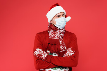Young man in santa hat, medical mask and mittens standing with crossed arms isolated on red