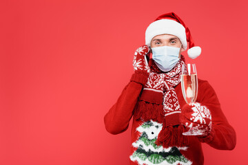 Man in santa hat and medical mask talking on smartphone while holding glass of champagne on blurred foreground isolated on red