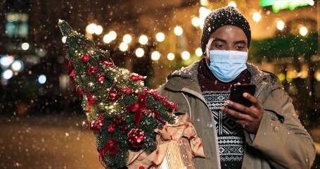 Cheerful man standing in snowy city and texting on cellphone while snowing. Young African American happy male tapping on smartphone outdoors with little Christmas tree Close up portrait Winter concept