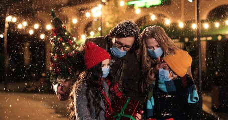 Portrait of happy family in masks standing outdoor while snowing and smiling. Joyful Caucasian boy and girl with parents on snowy street with presents. Quarantine. Holiday lights and gifts concept
