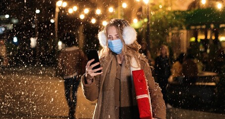 Obraz na płótnie Canvas Close up portrait of Caucasian young beautiful female in medical mask texting on smartphone while standing on street. Happy woman with christmas gift tapping on cellphone while snowing