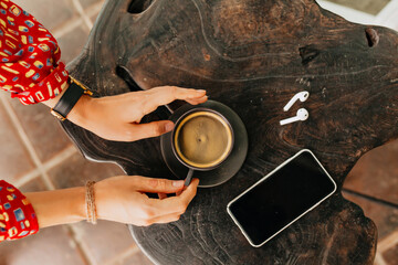 Fototapeta na wymiar Above frame of woman's hand holding a cup of coffee with headphones and smartphone over wood background