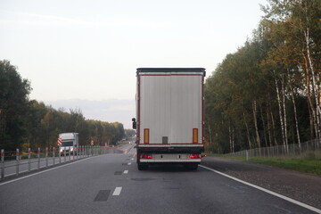 White van truck drive on two lane suburban asphalted highway road, back view at summer evening on forest and sky background, transportation logistics