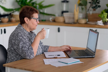 Senior woman using laptop for websurfing in her kitchen. The concept of senior employment, social security. Mature lady sitting at work typing a notebook computer in an home office.