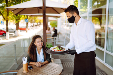 Young waiter in protective face mask and gloves while bringing food to a customer in cafe during coronavirus outbreak. Waiter serving in motion on duty in restaurant.