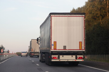 European road semi trucks with white trailer van move on two lane suburban asphalted highway motorway, rear view at summer evening on forest and sky background, delivery cargo logistics