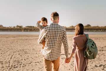 Happy family walking on sandy beach of river. Father, mother holding baby son on hands and going together. Rear view. Family Ties concept.