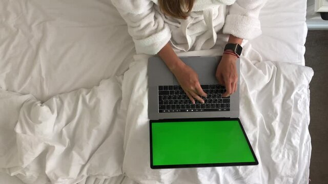 Woman working on macbook while lying in bed in hotel in a bathrobe and drinking coffee.